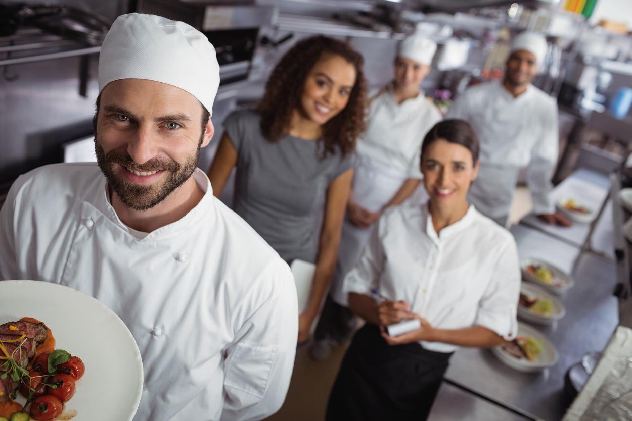 a chef wearing white clothes holding a vegetable plates with two other clefs and two waiters in the background